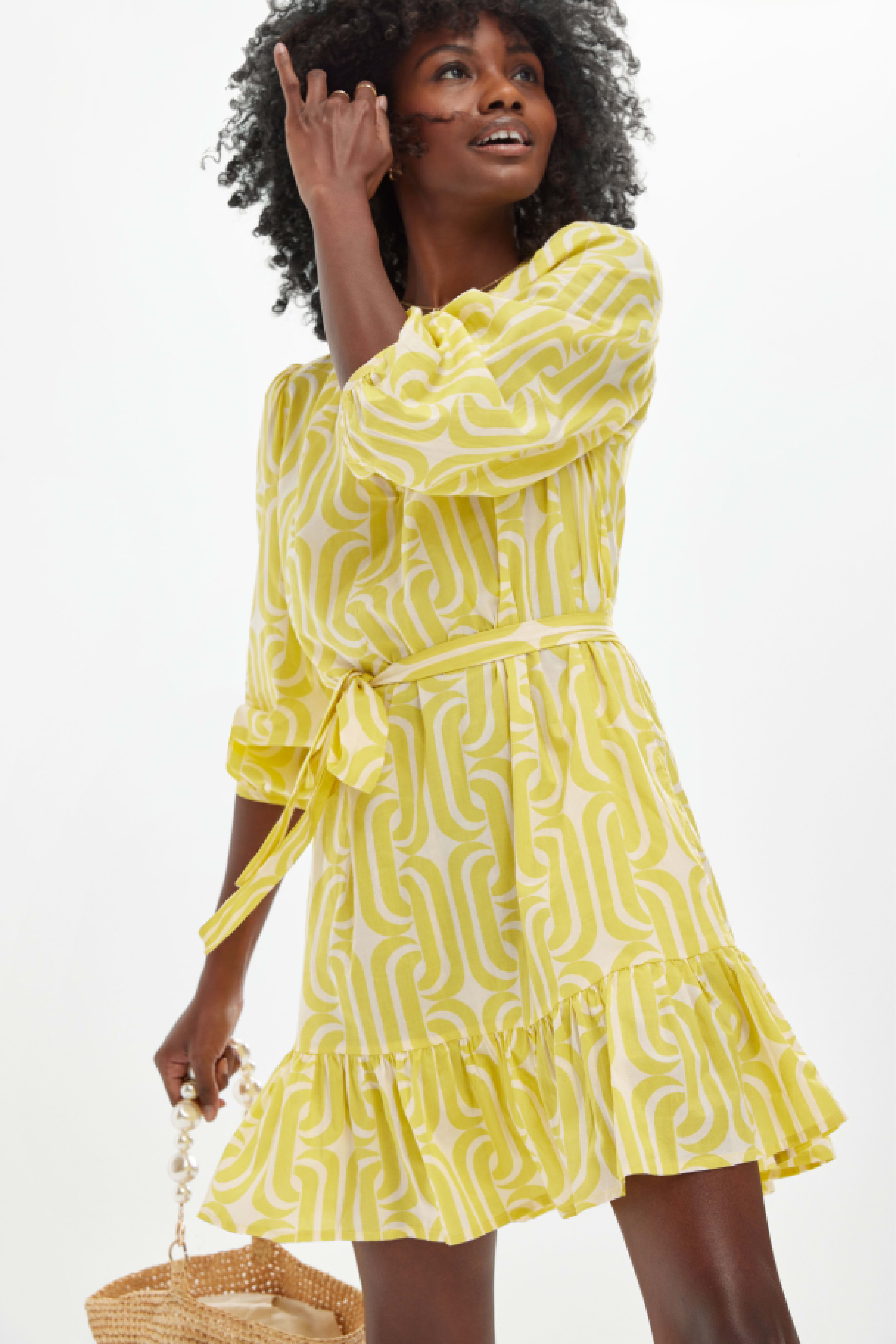 A woman in a yellow petite dress smiles and touches her hair.