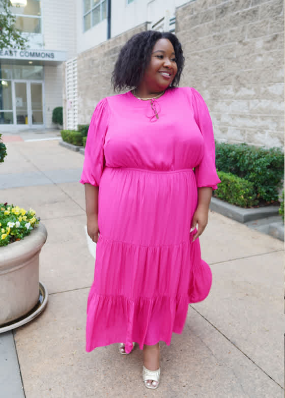 Model wearing Stitch Fix women’s clothing, including: a bright pink long-sleeve pleated midi dress with ruffle trim, paired with neutral heels.