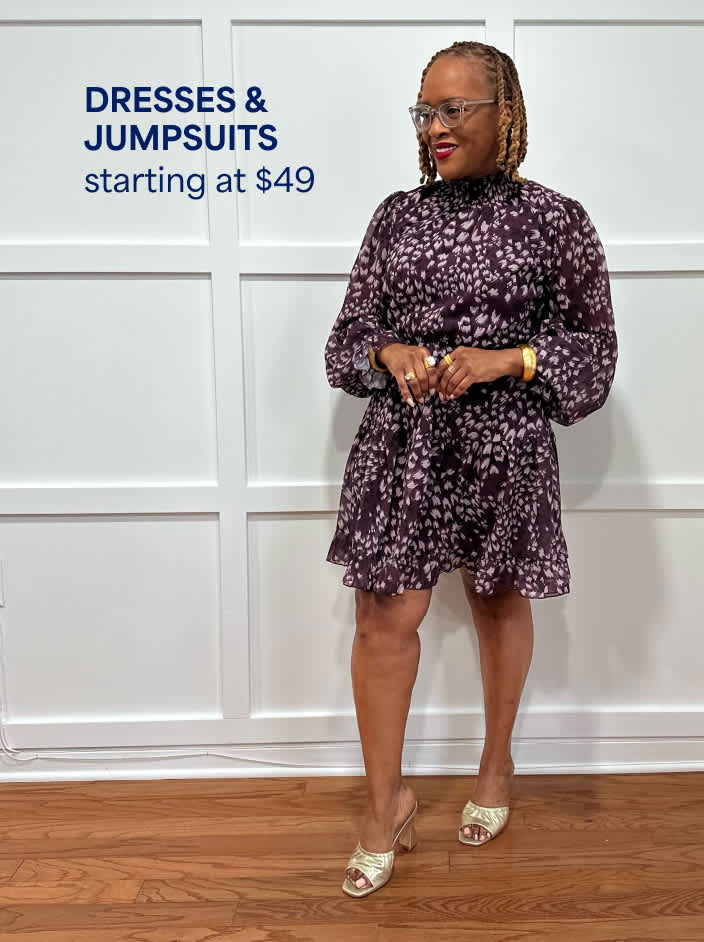 Dresses & Jumpsuits startingat $49. The image shows a petite woman in a black mini dress with a pink abstract pattern and open-toed heels.