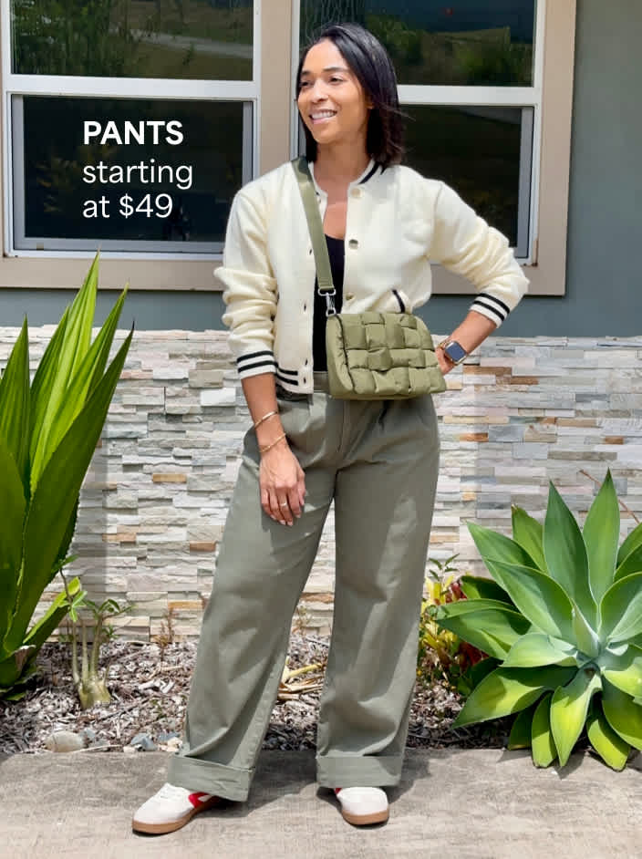 The image shows a petite woman in olive wide-leg pants, a cream varsity jacket, white sneakers and a green crossbody bag