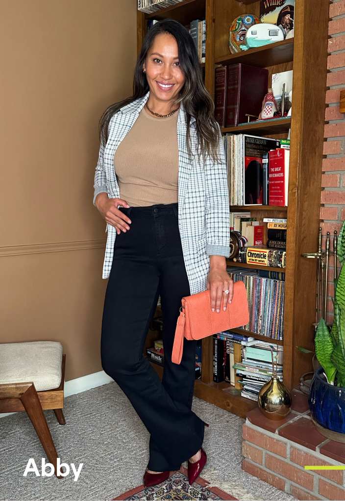 A petite woman with long brown hair wears a beige crewneck top with black pants, brown shoes, light blue and cream checked blazer and a coral clutch.