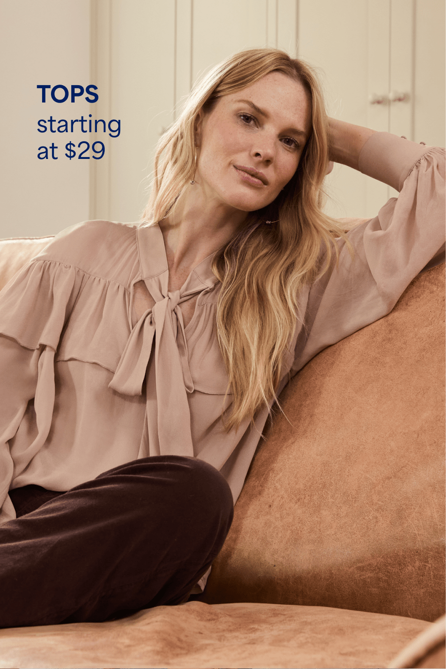 A woman with long blonde hair reclines on a couch, she wears a blush colored, flowy blouse with a bow at the neck and dark brown pants. Tops starting at $29