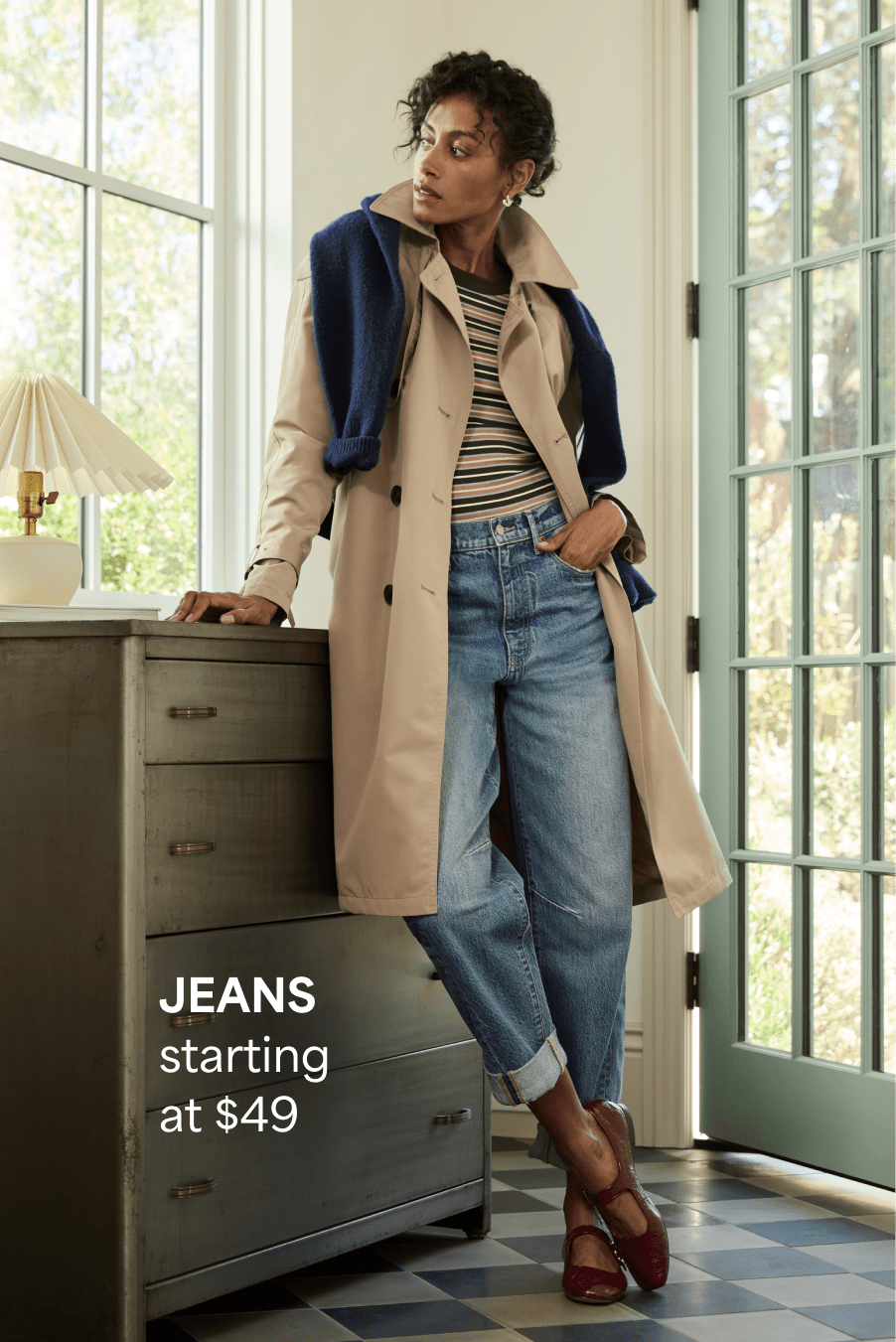 A woman with brown curly hair leans against a dresser, she wears cuffed light-wash jeans, a striped top and a trench coat with a dark blue sweater over her shoulders and burgundy Mary Janes. Jeans starting at $49 
