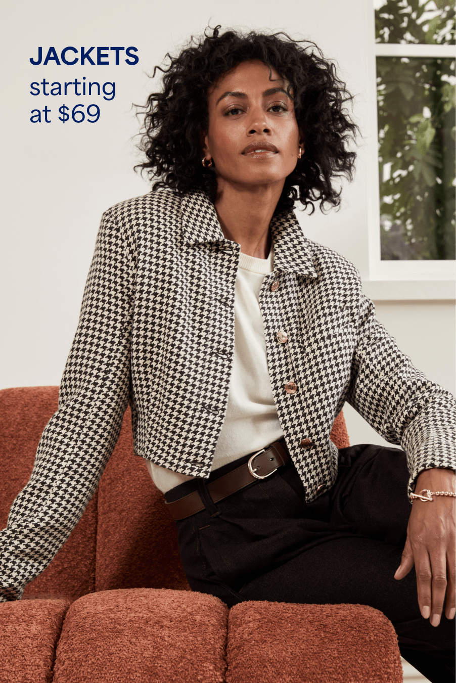 A woman with curly hair sits on the edge of a brown couch, she wears high-waisted dark trousers, a cream top and a cropped black-and-white houndstooth jacket. Jackets starting at $69