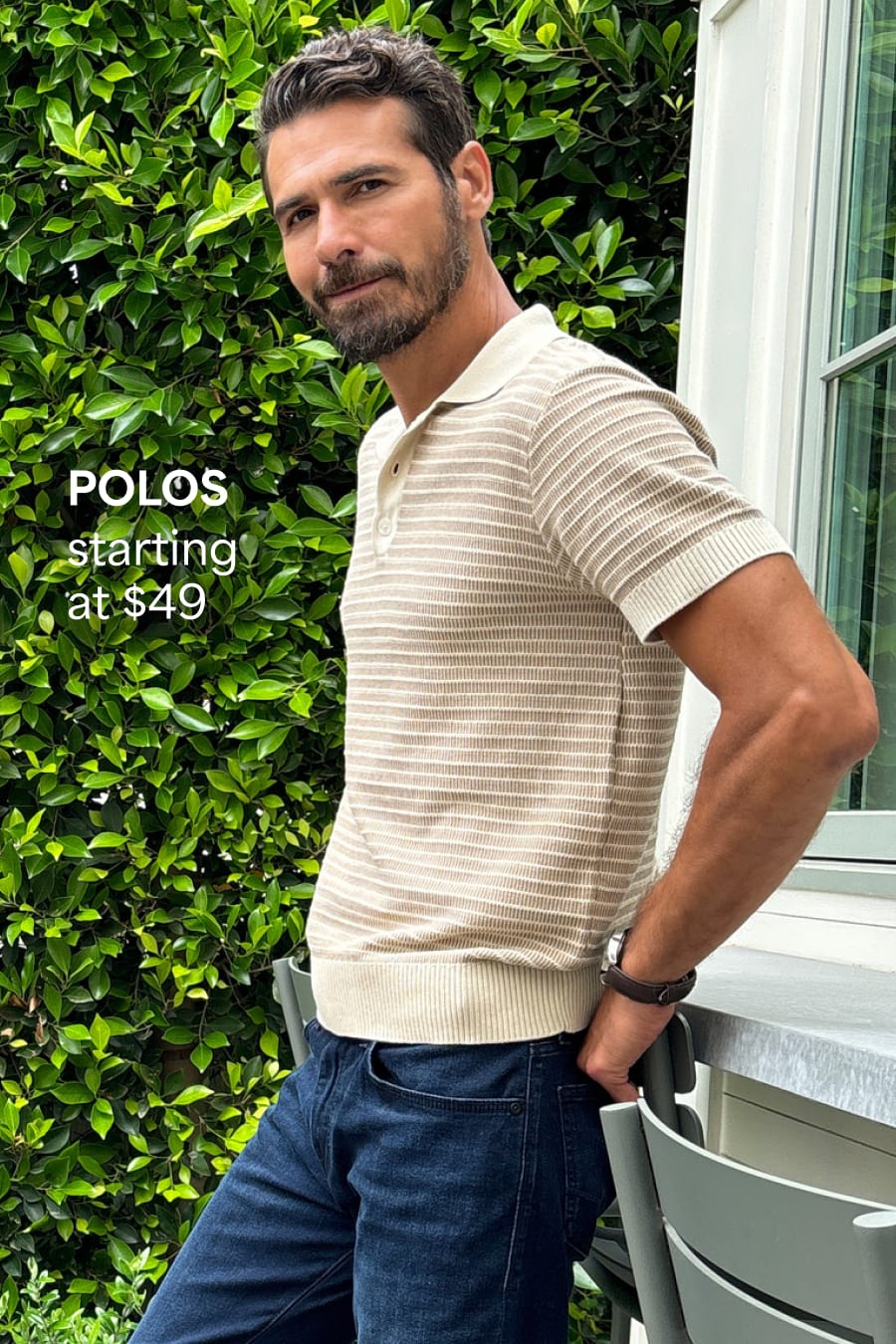 A bearded man wears a cream sweater polo with fine stripes and dark-wash jeans. Polos starting at $49