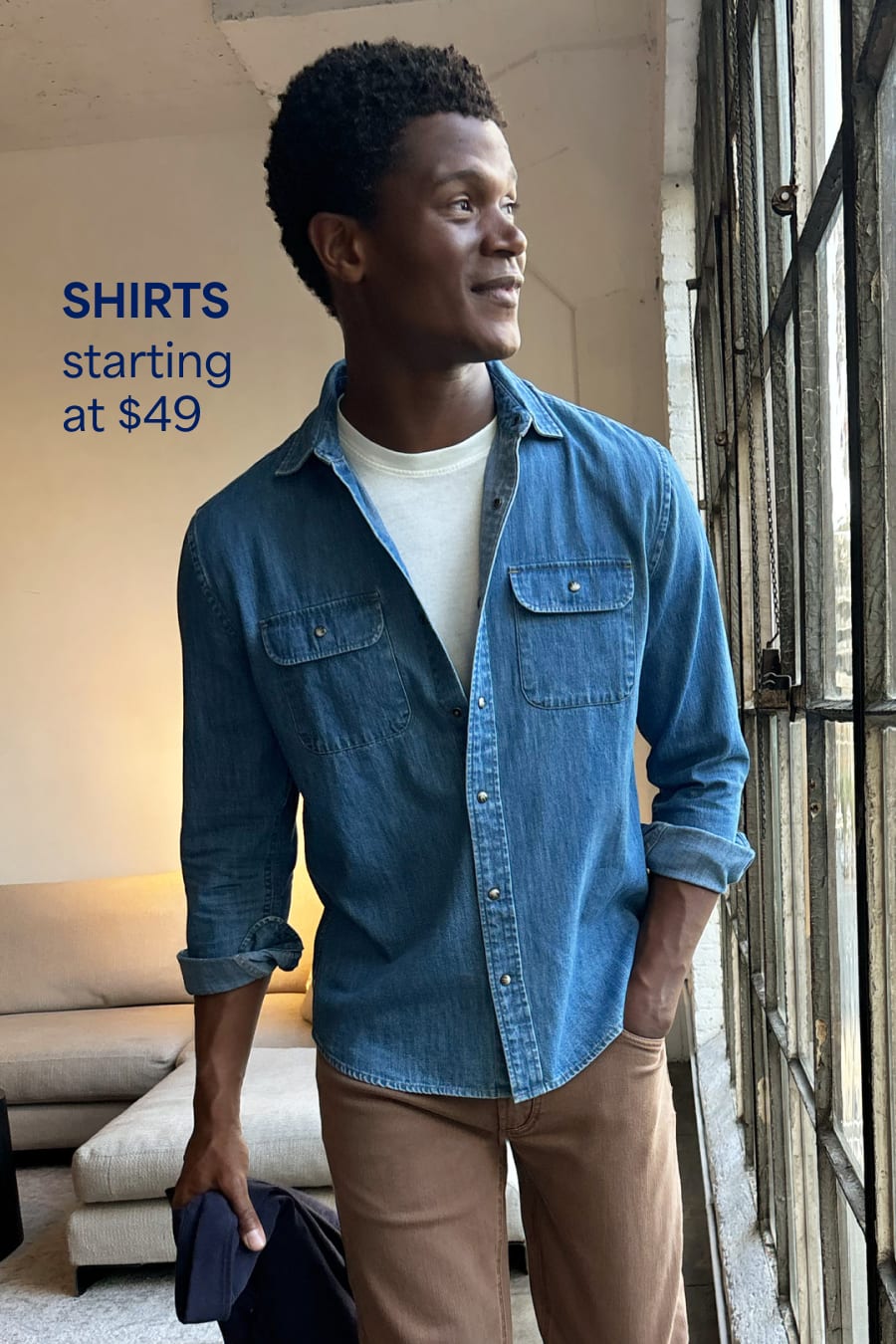 A man wears a denim shirt over a white t-shirt with brown jeans. Shirts starting at $49