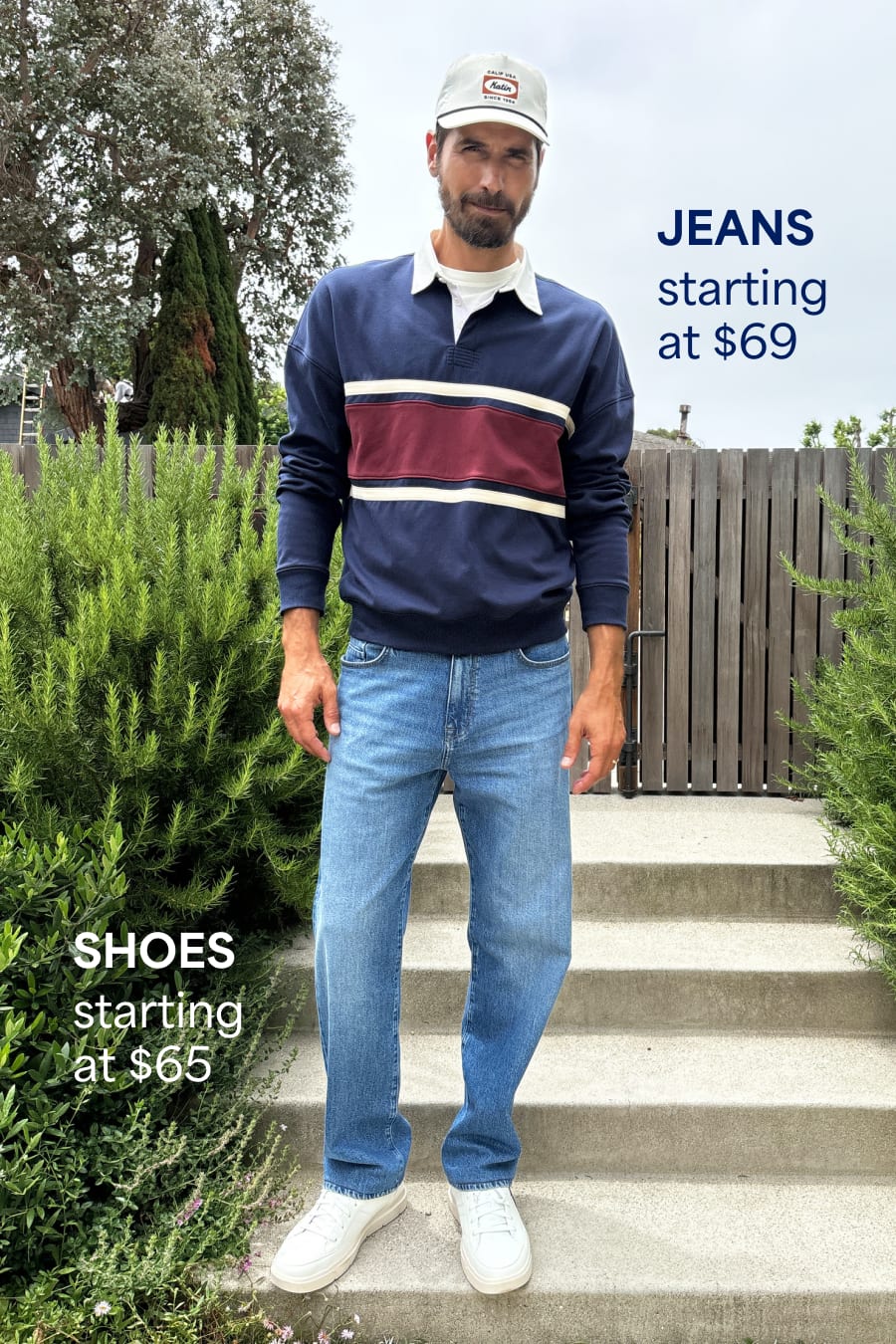 A bearded man wears a casual navy rugby-stripe sweatshirt, light blue jeans, white sneakers and a white cap. Jeans starting at $69. Shoes starting at $65