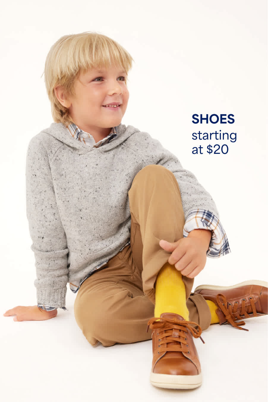 A boys wears a gray hoodie layered over a plaid shirt with tan pants, yellow socks and brown lace-up sneakers. Shoes starting at $20