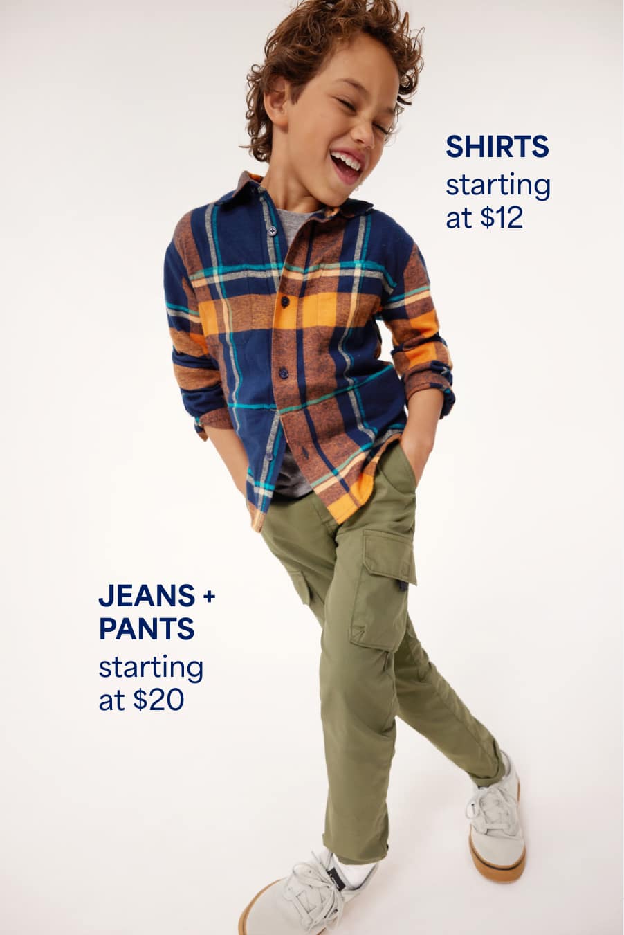 A boy wears a navy and orange plaid button-down over a gray tee with olive cargo pants. Shirts starting at $12, Jeans + pants starting at $20. 