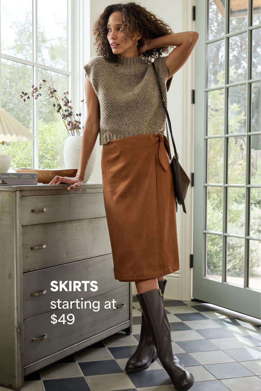 Stitch Fix women’s outfit featuring a woman in a sleeveless knit top, brown wrap midi skirt, and tall boots near a sunlit window. Text reads: “SKIRTS starting at $49.”
