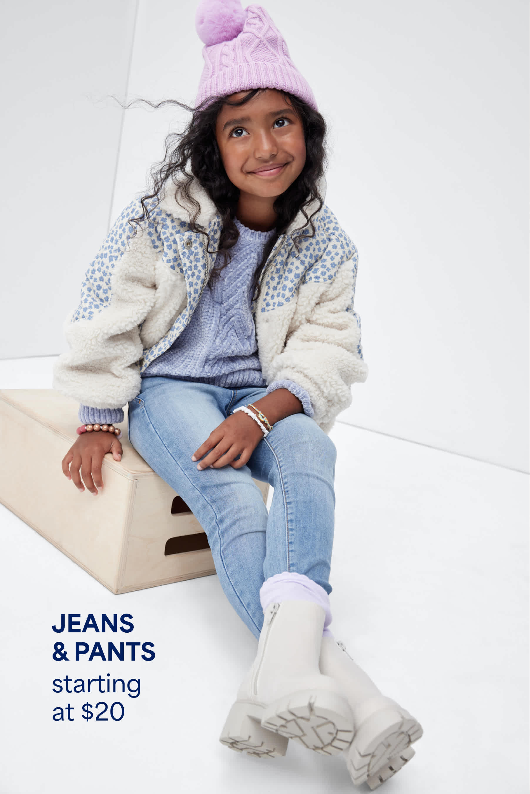 Stitch Fix Kids photo featuring a girl sitting and smiling, wearing a lavender knit sweater, white sherpa jacket, jeans, pink beanie, and white boots. Text reads: “JEANS & PANTS starting at $20.”
