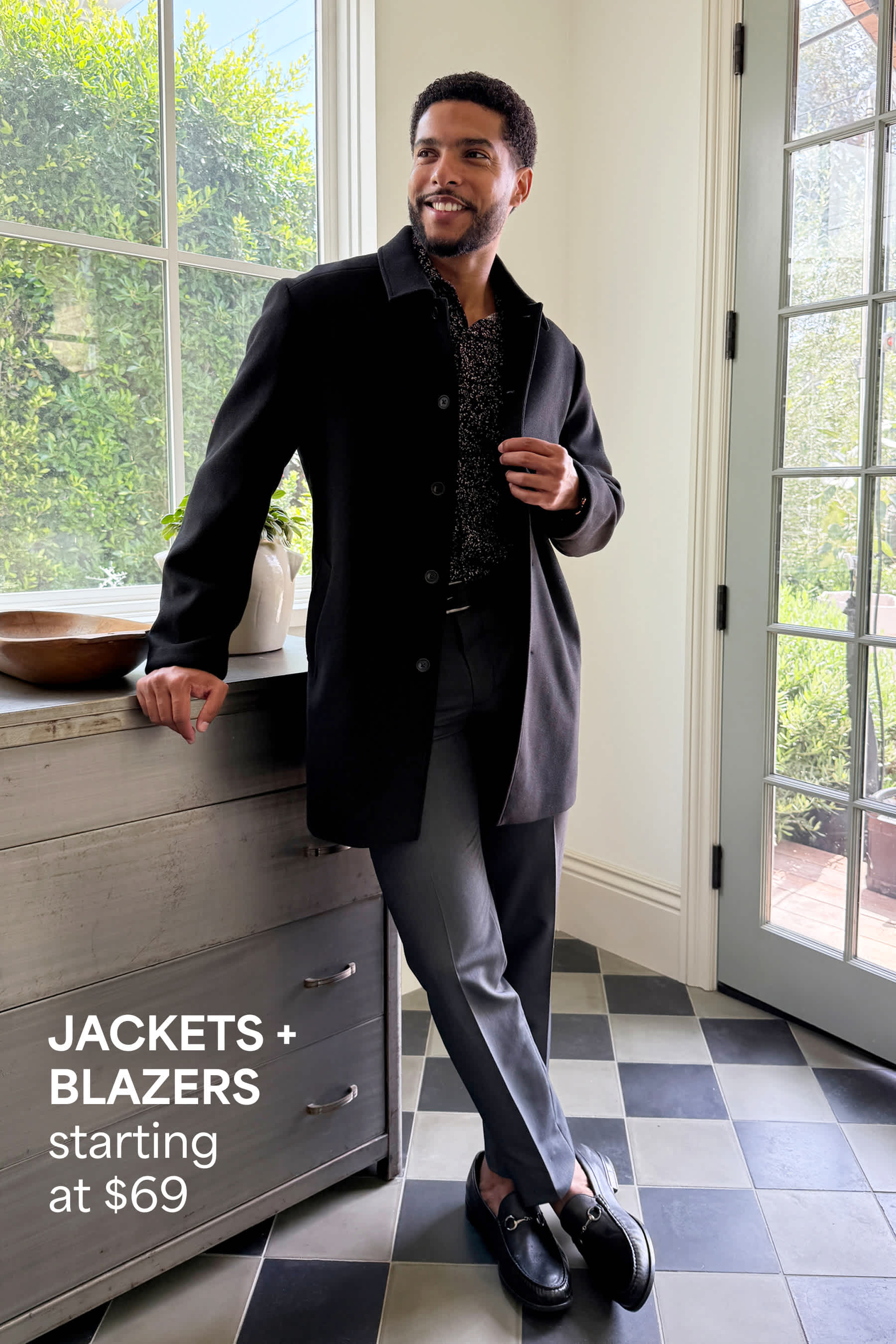 Stitch Fix men’s image showing a man in a black coat layered over a patterned shirt and dark trousers, standing near a sunlit entryway. Text reads: “JACKETS + BLAZERS starting at $69.”