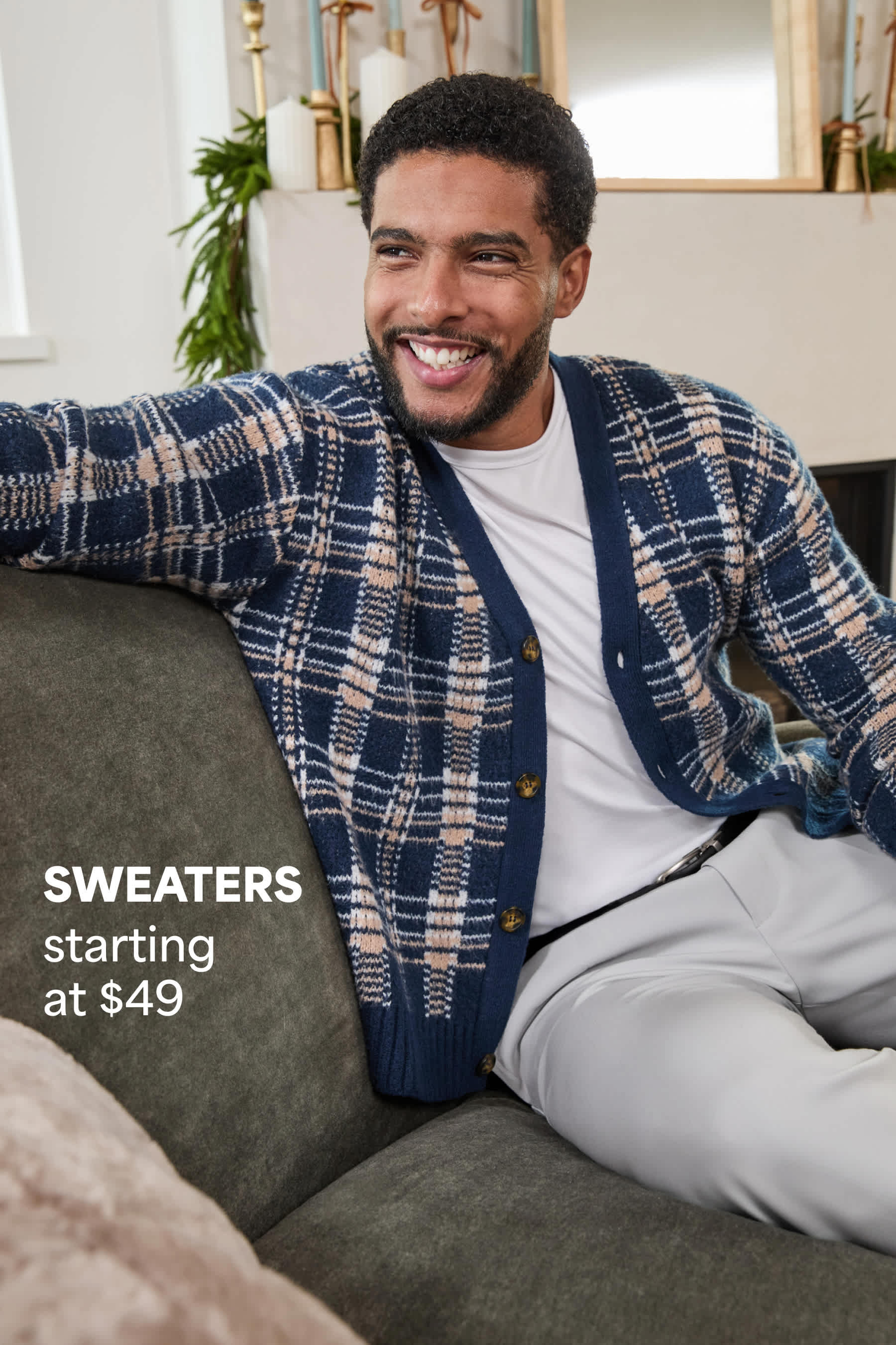 Stitch Fix men’s outfit featuring a man in a blue plaid cardigan layered over a white T-shirt and gray pants, sitting on a couch and smiling. Text reads: “SWEATERS starting at $49.”