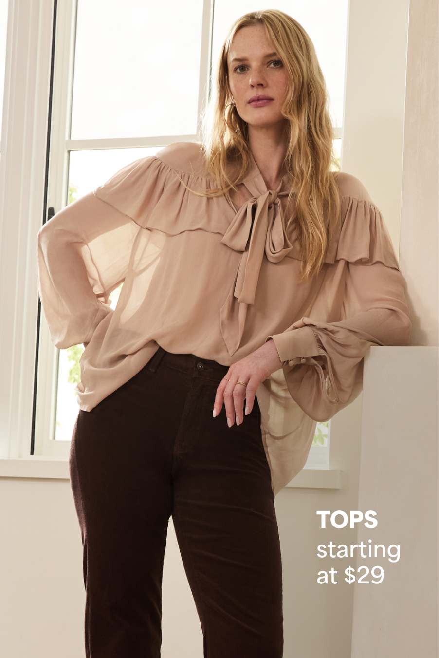 Stitch Fix women’s outfit featuring a woman in a beige bow-tie blouse and brown trousers, standing near a window. Text reads: “TOPS starting at $29.”