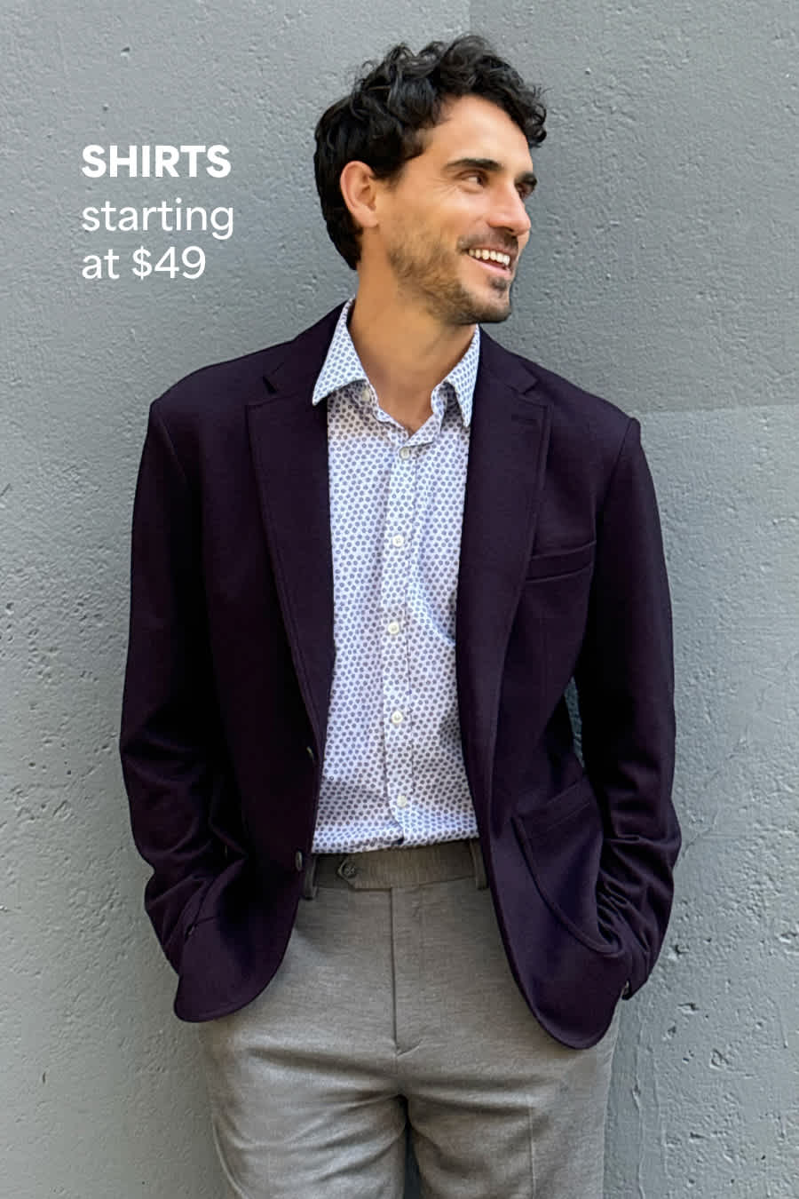 Stitch Fix male model posing against a wall wearing a printed button-down shirt, plum blazer and gray trousers. Text overlay on image reads “Shirts starting at $49”