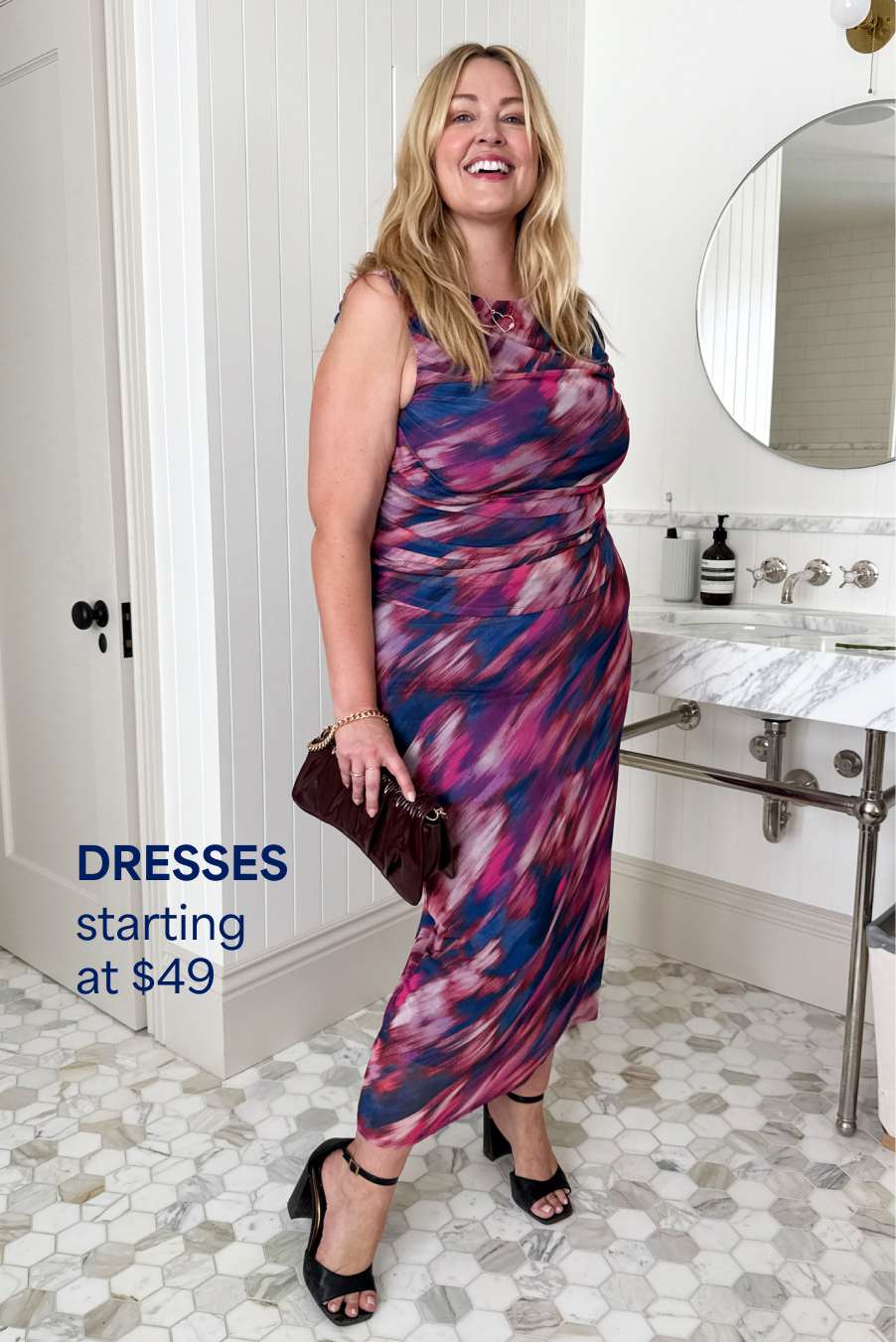 Stitch Fix woman model posing in front of a bathroom vanity wearing a printed pink and purple dress, black heels and a clutch. Text overlay on the image reads “Dresses starting at $49”