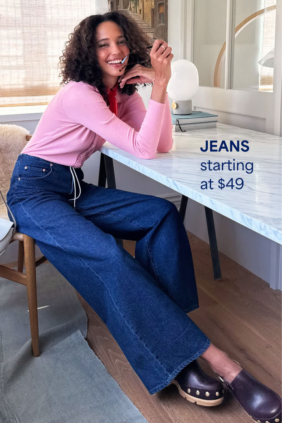 Stitch Fix woman model posing and sitting down at a desk wearing a pink cardigan, dark-wash jeans and studded clogs. Text overlay on image reads “Jeans starting at $49”