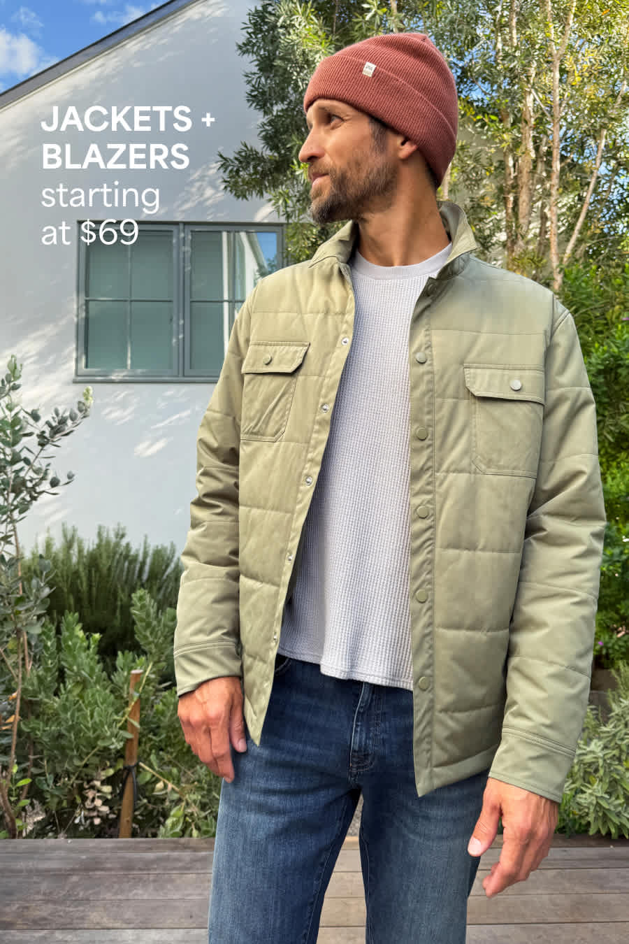 Stitch Fix male model posing outdoors in front of a house wearing a rust-colored beanie, gray waffle shirt, green quilted jacket and dark-wash jeans. Text overlay on image reads “Jackets + blazers starting at $69”