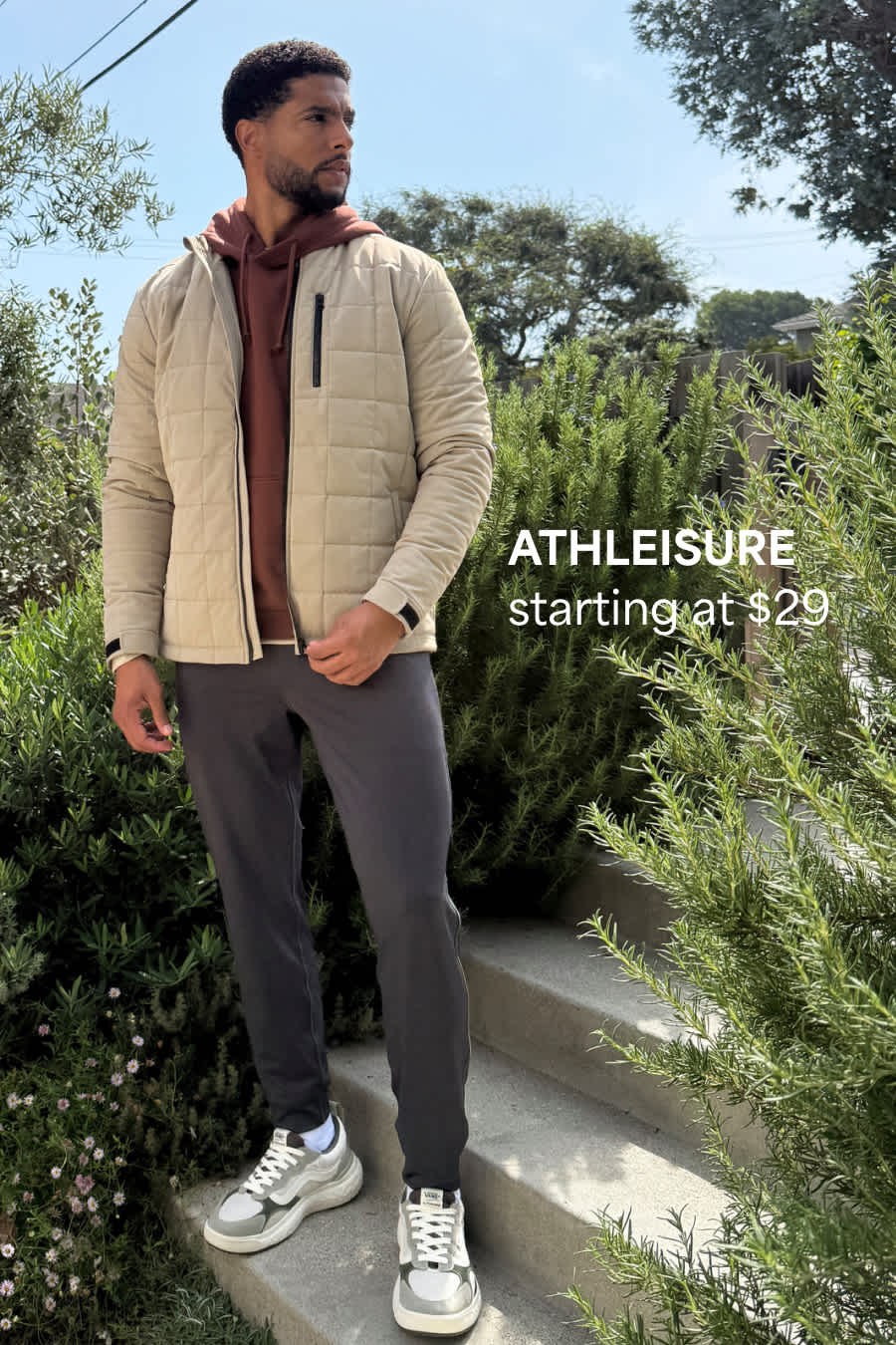 Stitch Fix male model posing outdoors wearing a brown hoodie, cream quilted jacket, gray joggers and active sneakers. Text overlay on image reads “Athleisure starting at $29”