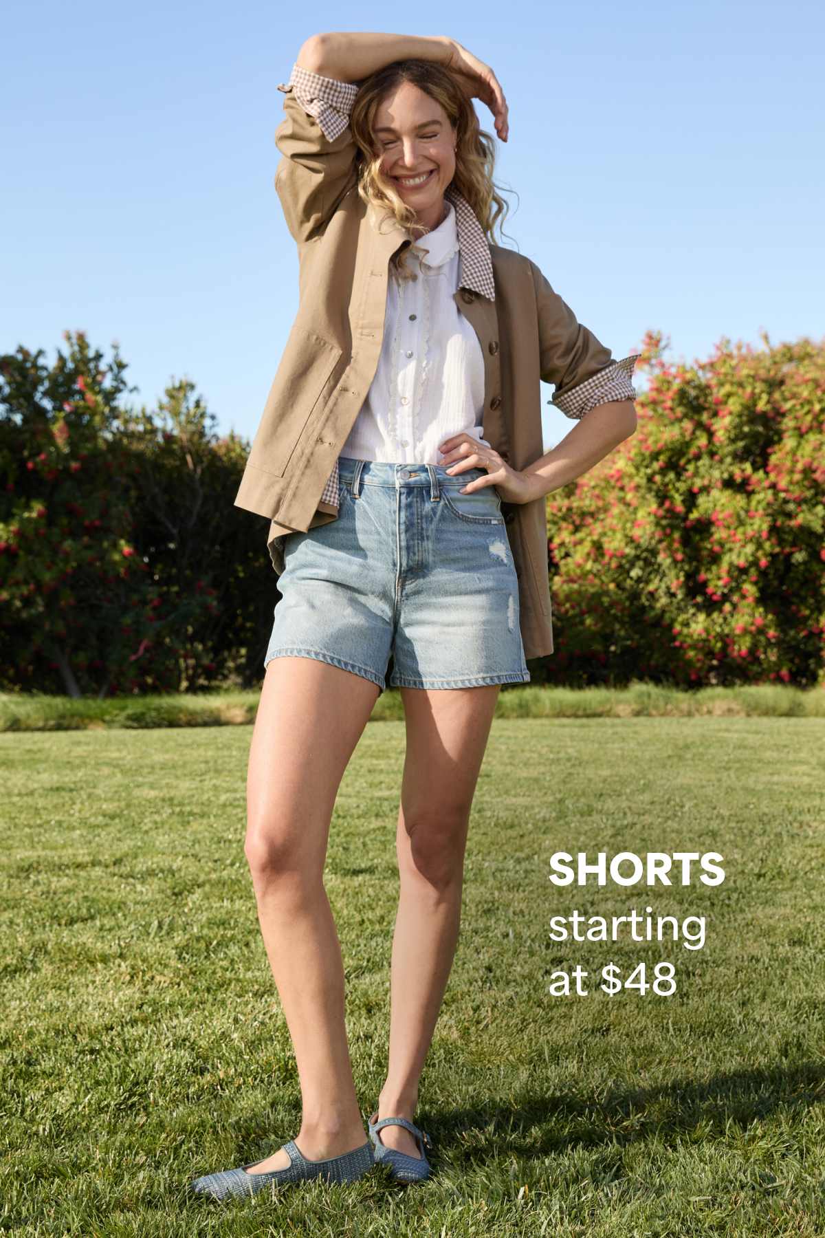 Woman standing on grass wearing a casual outfit with a light jacket and denim shorts. Text reads “SHORTS starting at $48.”