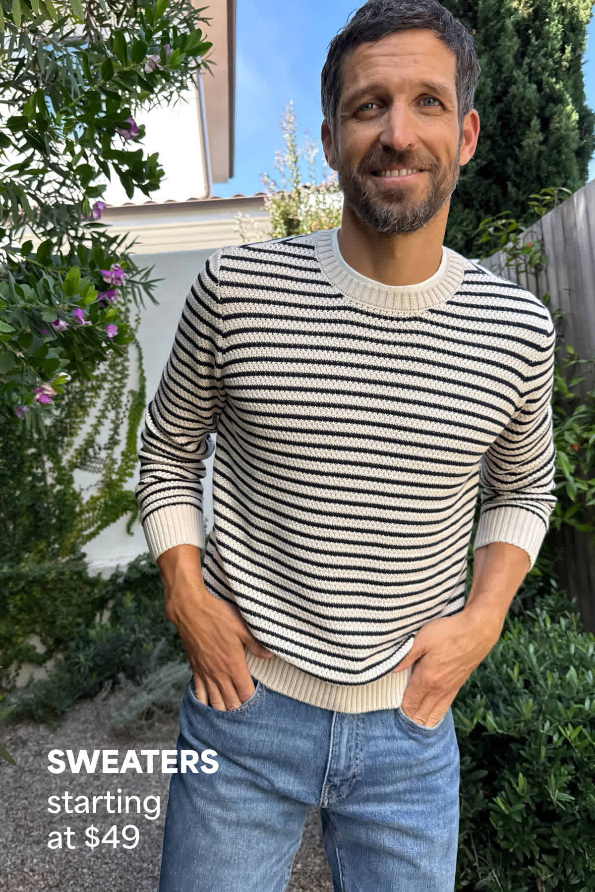 A man stands outdoors in a garden setting wearing a striped crewneck sweater and blue jeans, with his hands in his pockets and a relaxed smile. Text on the image reads “Sweaters starting at $49,” highlighting the starting price for sweaters.