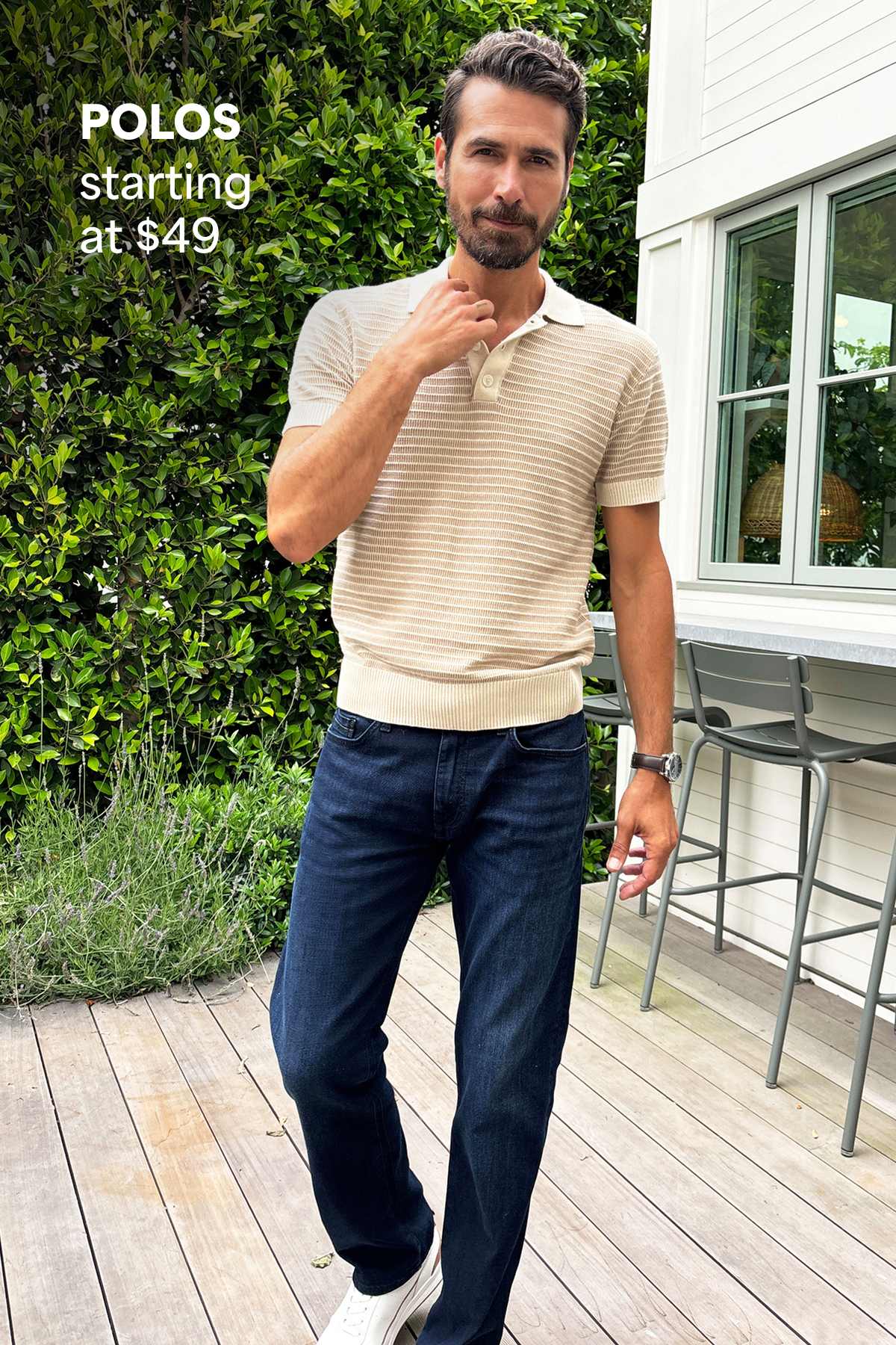A man stands on a wooden deck outdoors wearing a short-sleeve knit polo shirt and dark jeans, with greenery and a home exterior behind him. Text on the image reads “Polos starting at $49,” highlighting the starting price for polos.