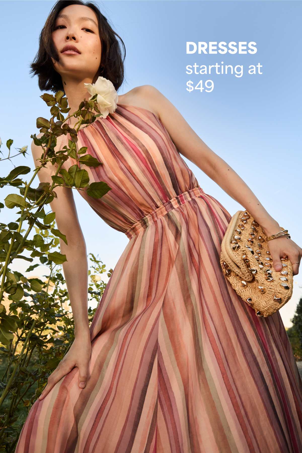 Woman outdoors wearing a flowy striped dress and holding a woven clutch. Text reads “DRESSES starting at $49.”