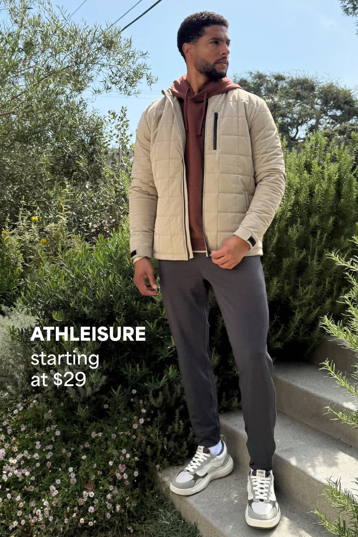 A man stands outdoors on steps surrounded by greenery, wearing an athleisure outfit with a light quilted jacket layered over a hoodie, jogger-style pants, and sneakers. Text on the image reads “Athleisure starting at $29,” highlighting the starting price for athleisure styles.