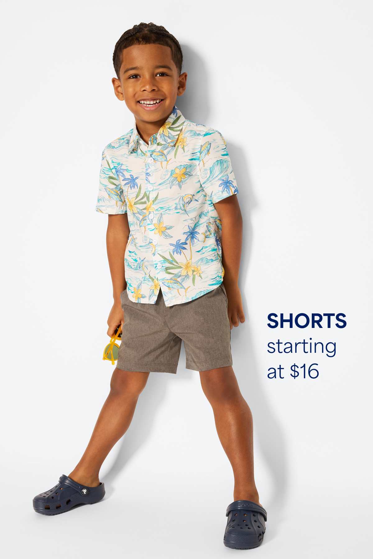 A smiling boy stands against a light background wearing an open button-up shirt layered over a graphic T-shirt, paired with jeans and sneakers. Text on the image reads “Jeans + Pants starting at $20,” highlighting the starting price for kids’ jeans and pants.