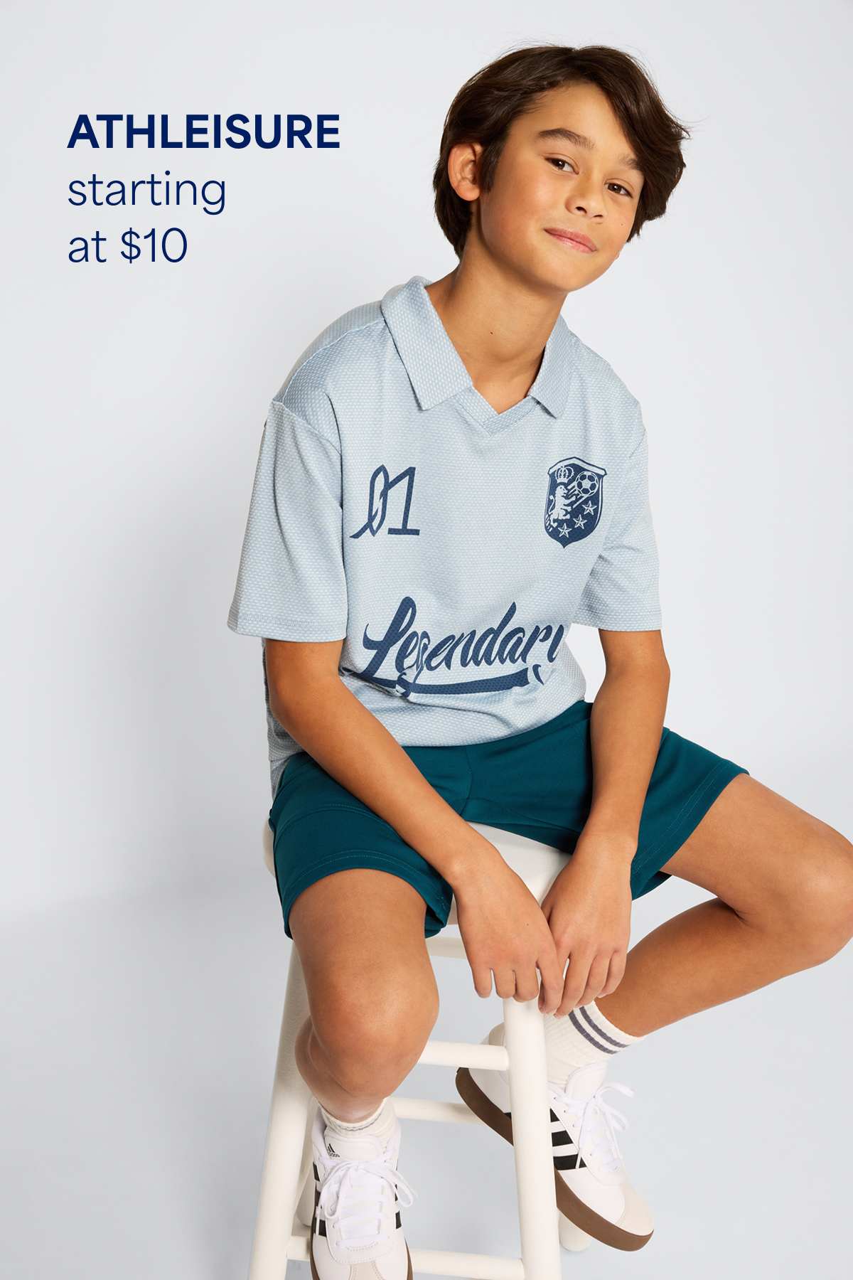 A boy sits on a stool against a light background wearing an athleisure outfit with a short-sleeve sports-style top, athletic shorts, and sneakers. Text on the image reads “Athleisure starting at $10,” highlighting the starting price for kids’ athleisure styles.