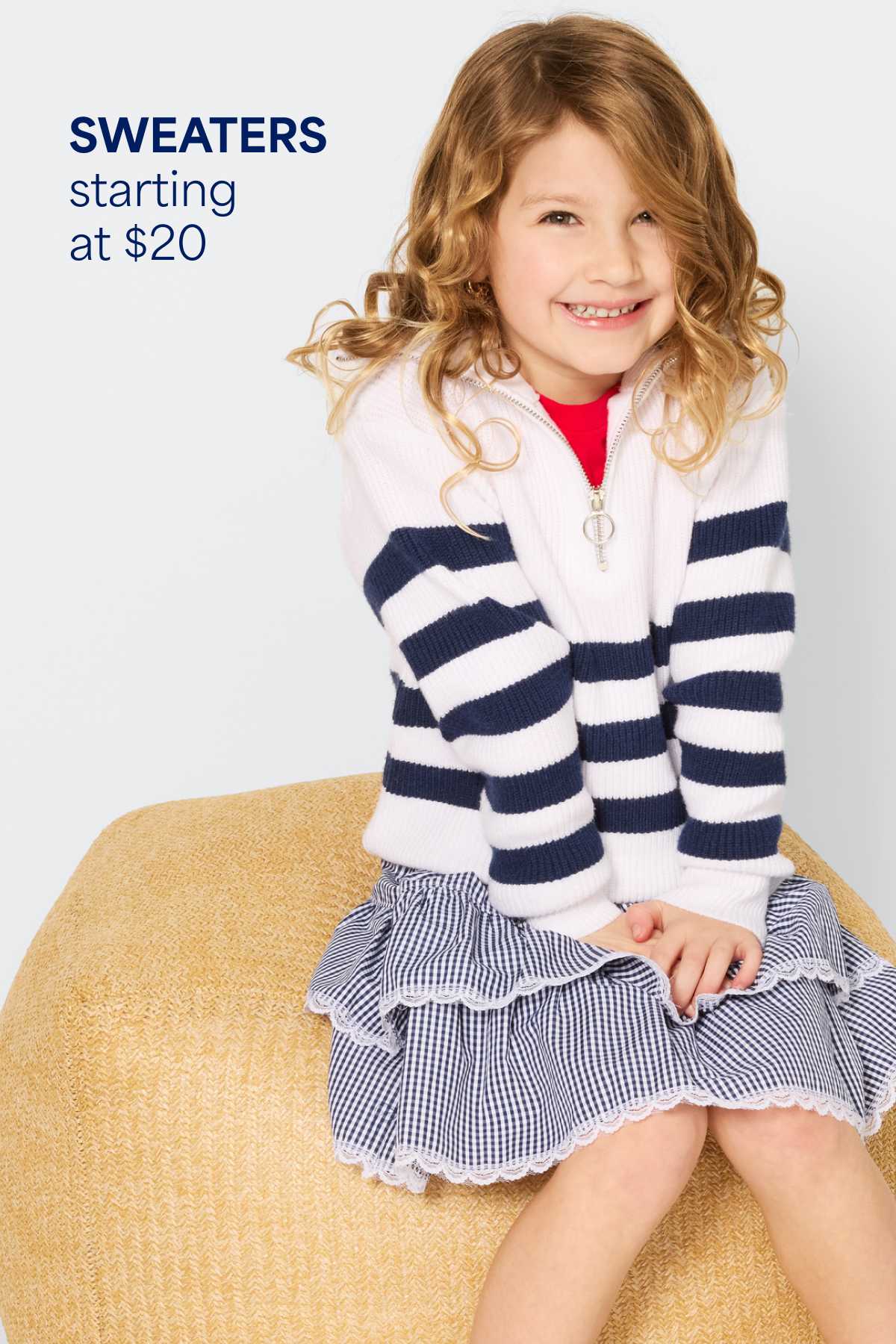 A smiling girl kneels against a light background wearing a bright yellow sweatshirt, light-wash cargo-style jeans, and a pink sweater tied around the waist. Text on the image reads “Tops starting at $10,” highlighting the starting price for kids’ tops.