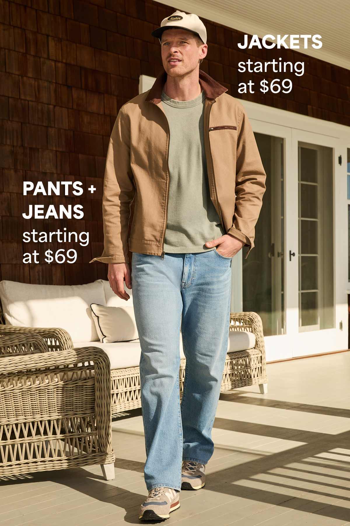 A man walks on an outdoor porch wearing a tan jacket layered over a green T-shirt, light-wash jeans, sneakers, and a baseball cap. Text on the image reads “Jackets starting at $69” and “Pants + Jeans starting at $69,” highlighting starting prices for the featured categories.