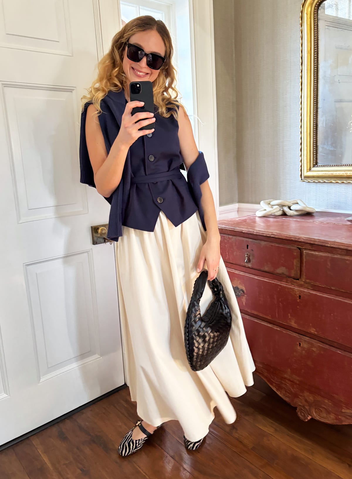 Woman in navy vest, cream midi skirt, black flats, carrying black woven bag