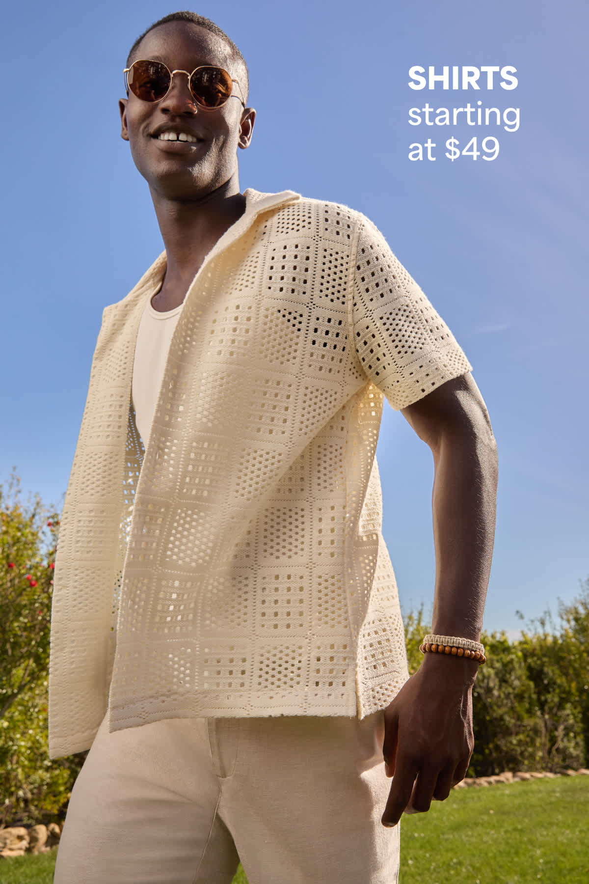 A man models a cream open-knit short-sleeve shirt and light neutral pants outdoors beside text that reads “SHIRTS starting at $49.”