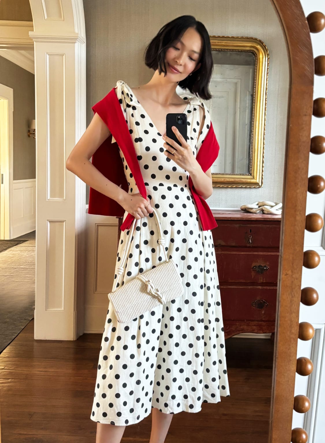 Woman in white polka dot midi dress with red cardigan taking mirror selfie