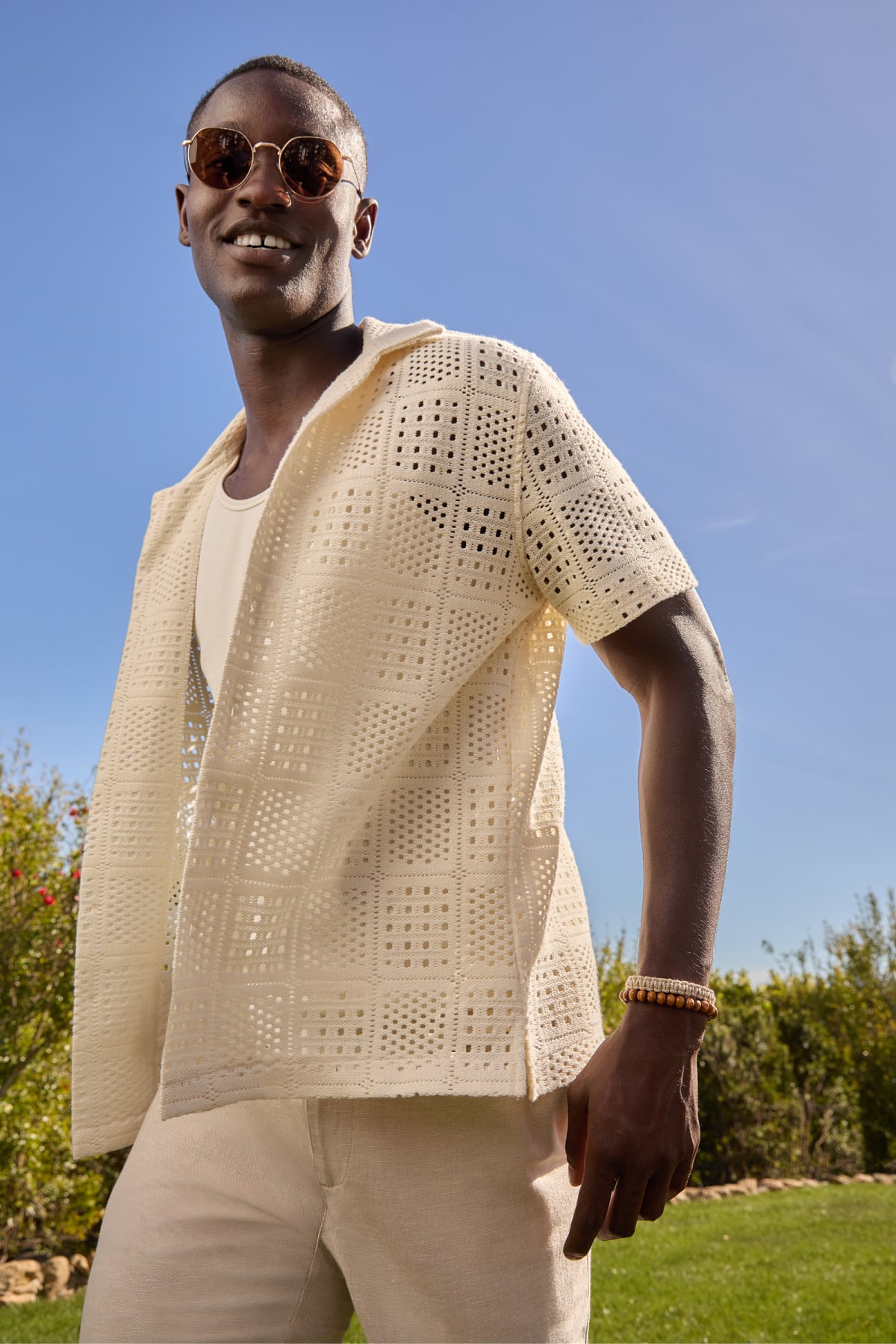 Model wearing Stitch Fix men’s clothes including a cream crochet short-sleeved shirt, white tank top, and light khaki pants.