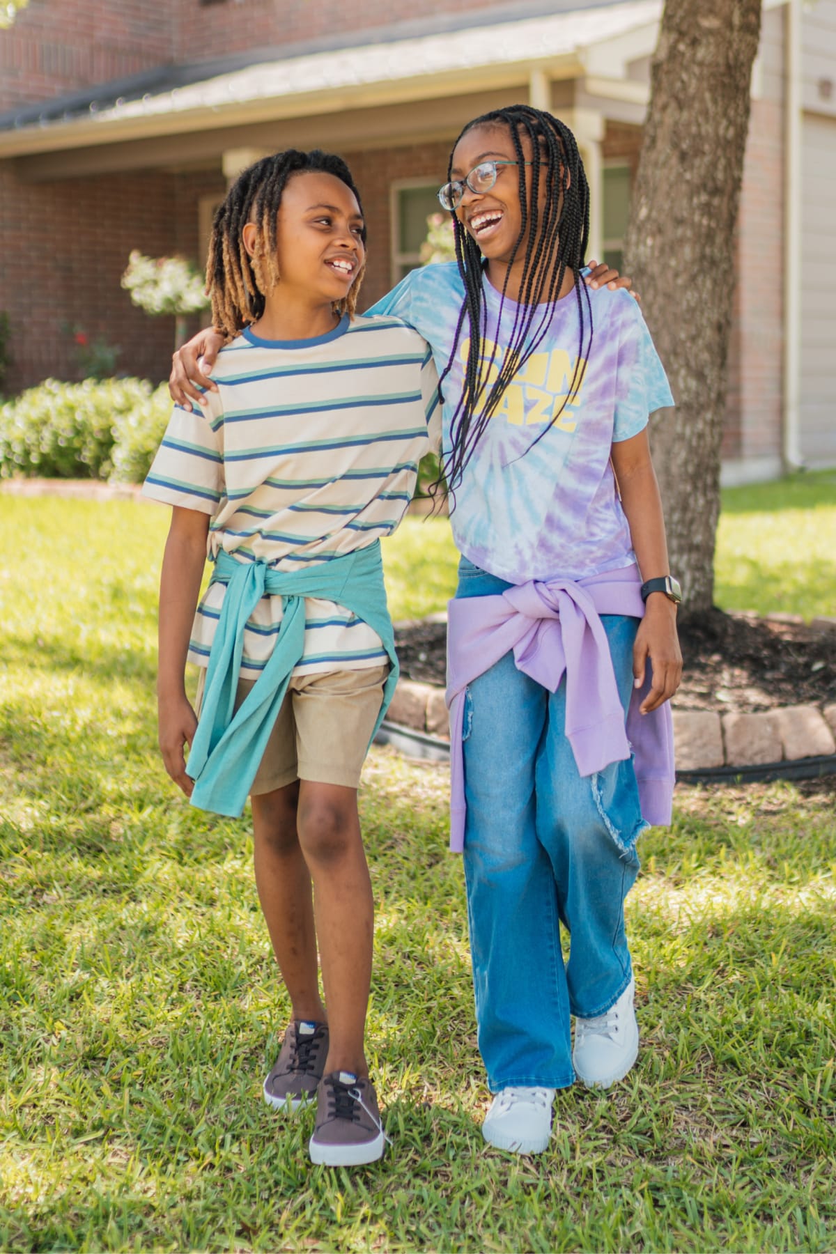 Two kids wearing Stitch Fix clothes including a tie-dye graphic tee, blue and beige striped shirt, light khaki shorts, denim shorts, and sneakers.