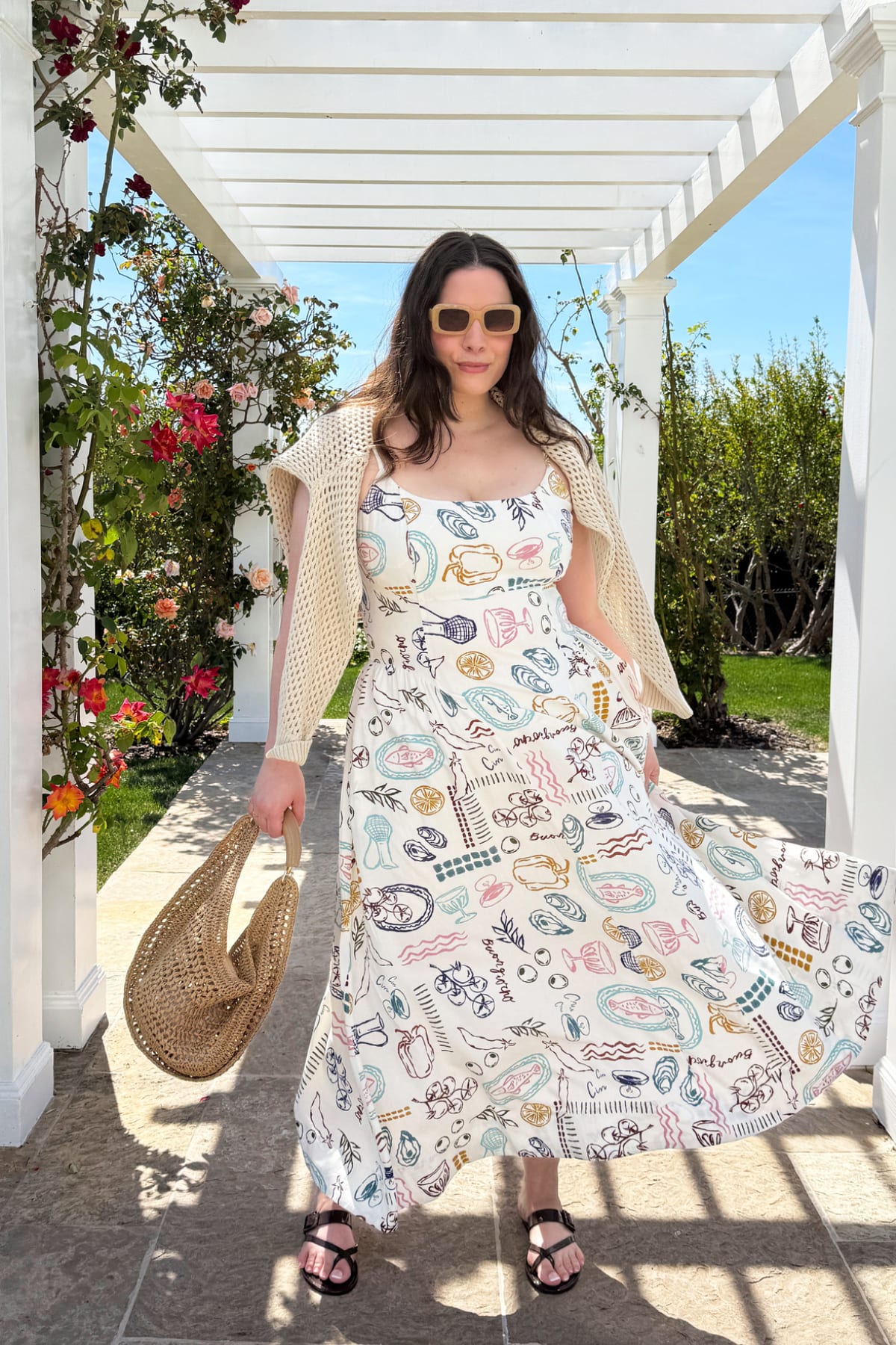 Model wearing Stitch Fix women’s clothes including a printed white maxi dress, cream open-knit sweater, black sandals, and carrying a woven bag.