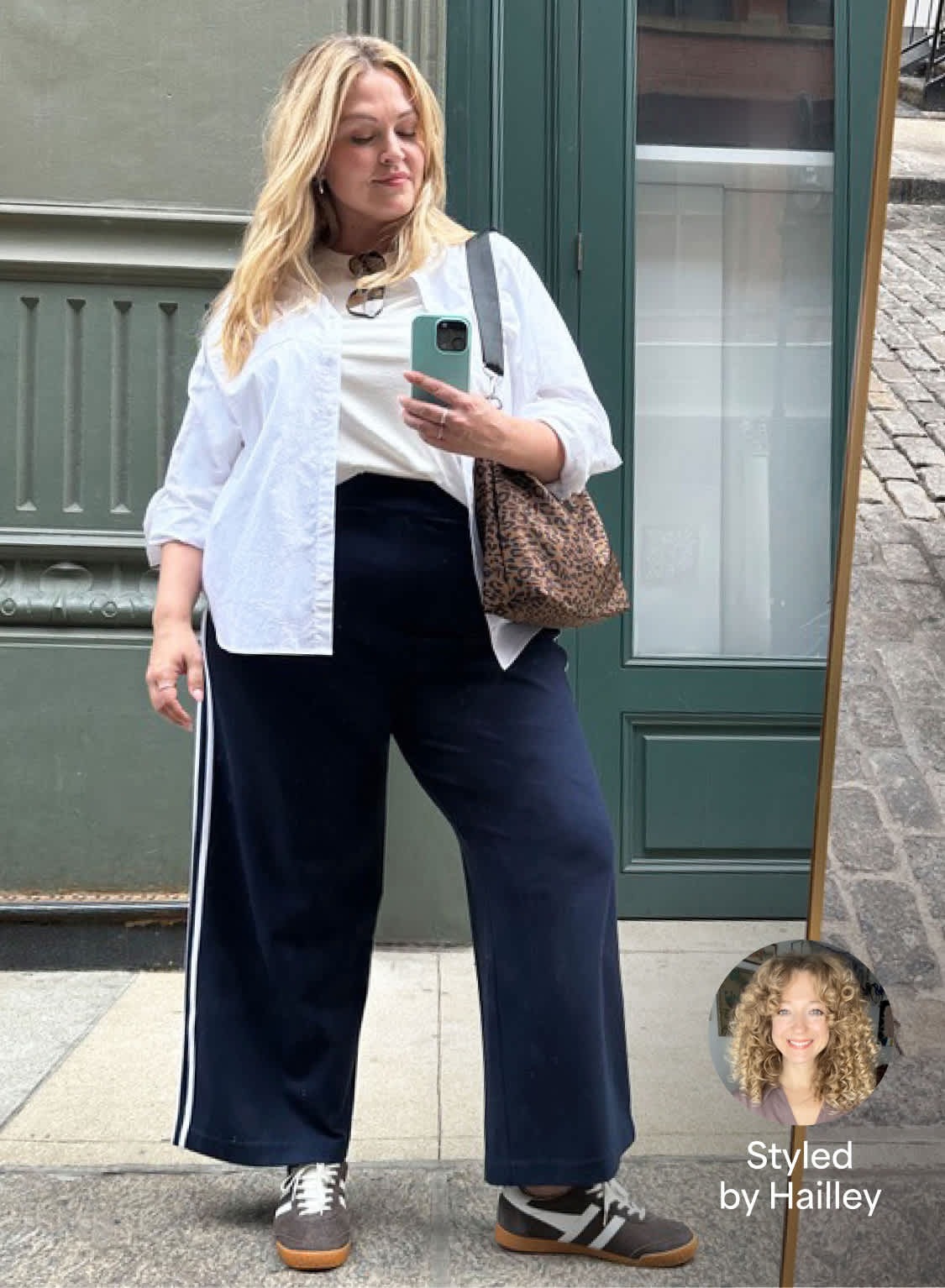 A person models a casual outfit featuring navy wide-leg track pants with white side stripes, paired with a white crewneck tee, an open white button-up shirt, gray suede sneakers, and a leopard-print shoulder bag. A product cutout of the navy pants appears in the upper left, while a small circular "Styled by Hailley" badge with a stylist headshot is shown in the lower right. The look is photographed in a full-length mirror outdoors in front of a green storefront.