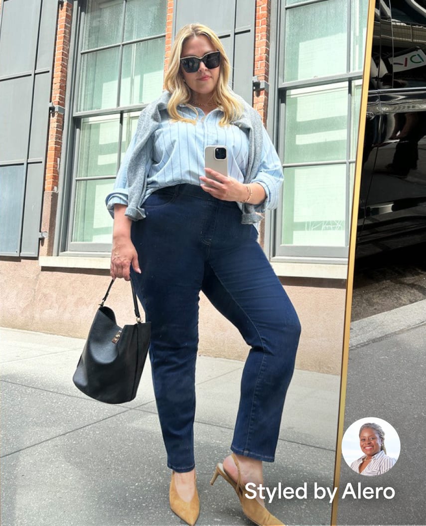 Woman wearing a blue pinstripe shirt, grey sweater, dark blue jeans, brown heels, carrying a black tote bag. Styled by Alero