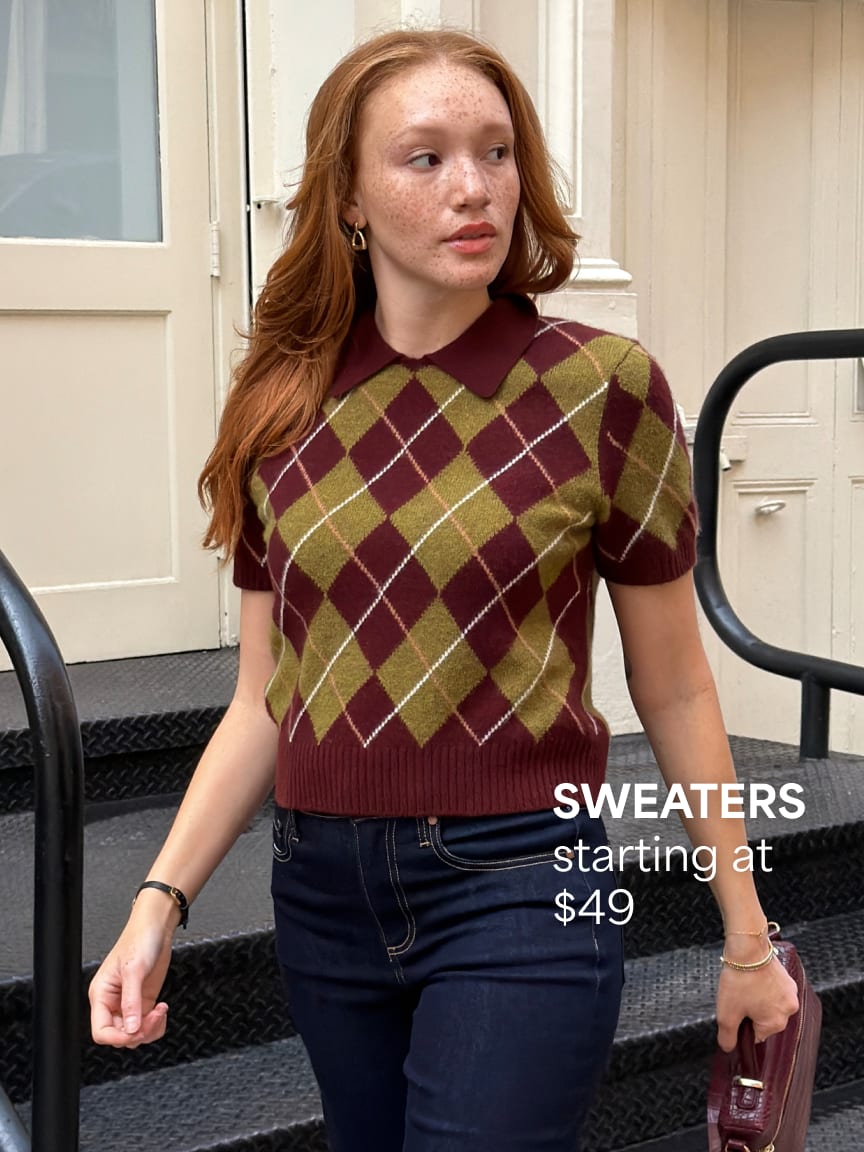 Woman wearing a short-sleeve maroon and olive green argyle collared sweater, dark blue jeans, carrying a bag. Sweaters starting at $49