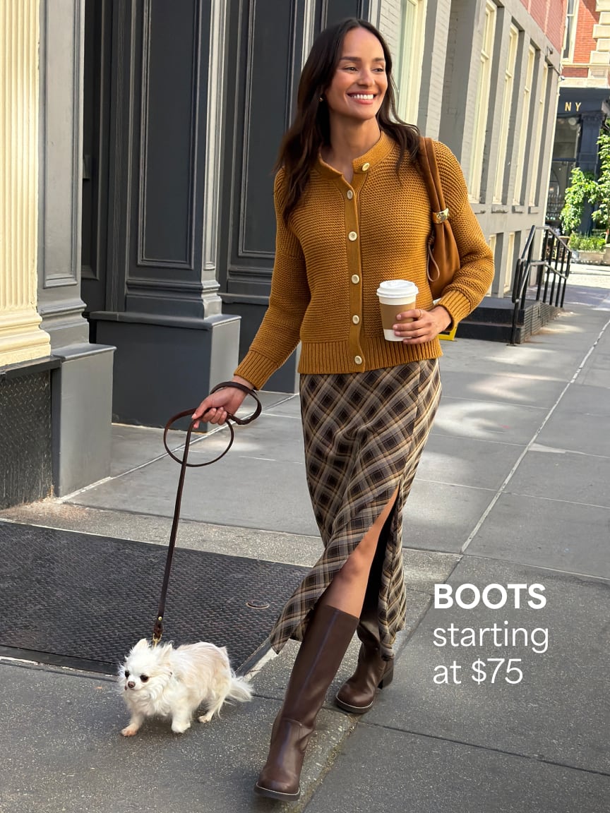 Woman wearing a mustard yellow cardigan sweater, plaid slit skirt, dark brown knee-high boots, walking a small dog. Boots starting at $75