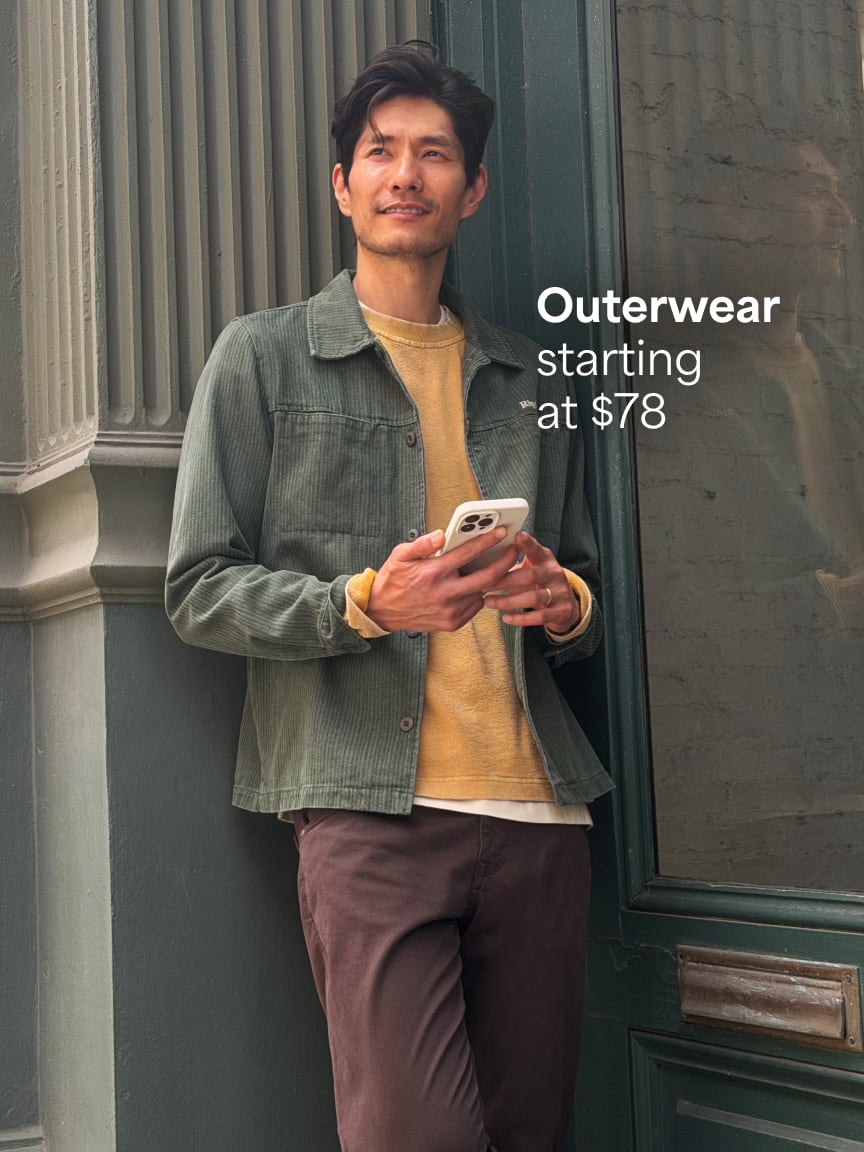 Man in green jacket, yellow sweater, white t-shirt, and brown pants looking at a smartphone. Text: Outerwear starting at $78