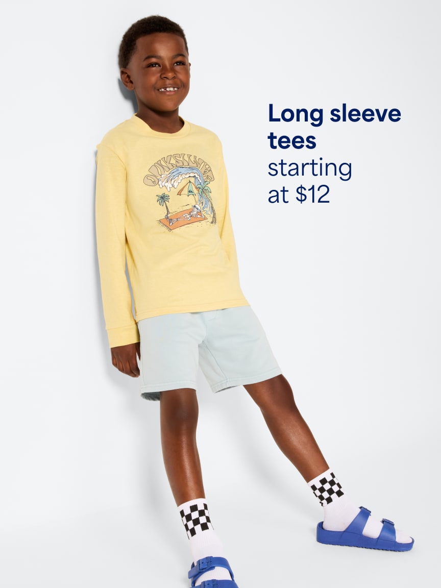 Child wearing a yellow long-sleeve graphic tee, light blue shorts, checkered socks, and blue sandals. Text: Long-sleeve tees starting at $12
