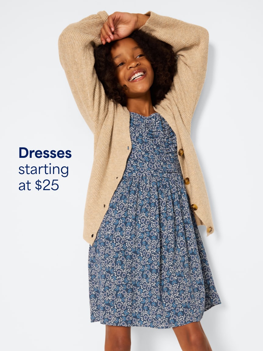 Girl wearing a blue floral print dress layered under a tan knit cardigan sweater. Text: dresses starting at $25