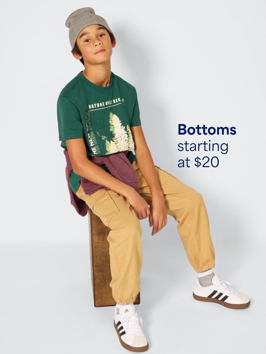 Boy wearing a grey beanie, green graphic t-shirt, tan cargo pants, white sneakers, and maroon sweater tied around waist. Text: Bottoms starting at $20