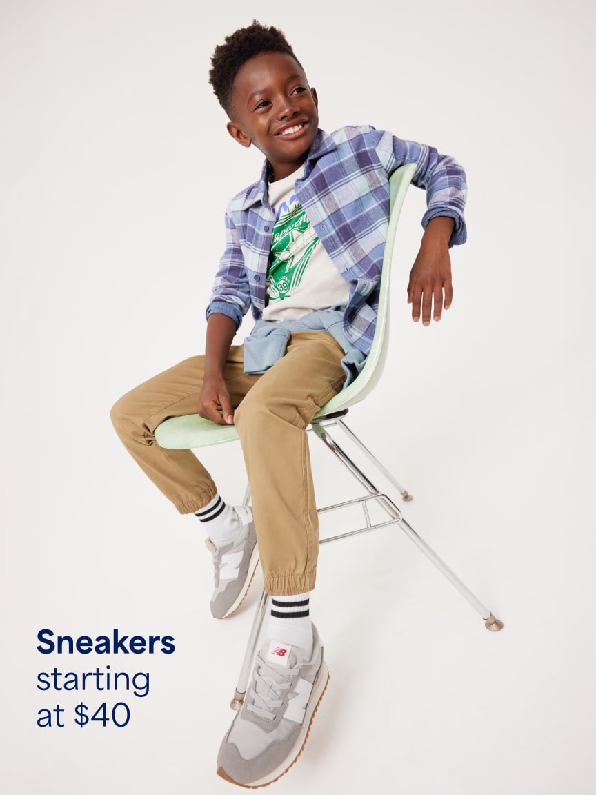 Boy sitting on a chair wearing a blue plaid shirt over a graphic tee, tan pants, and grey sneakers. Text: Sneakers starting at $40