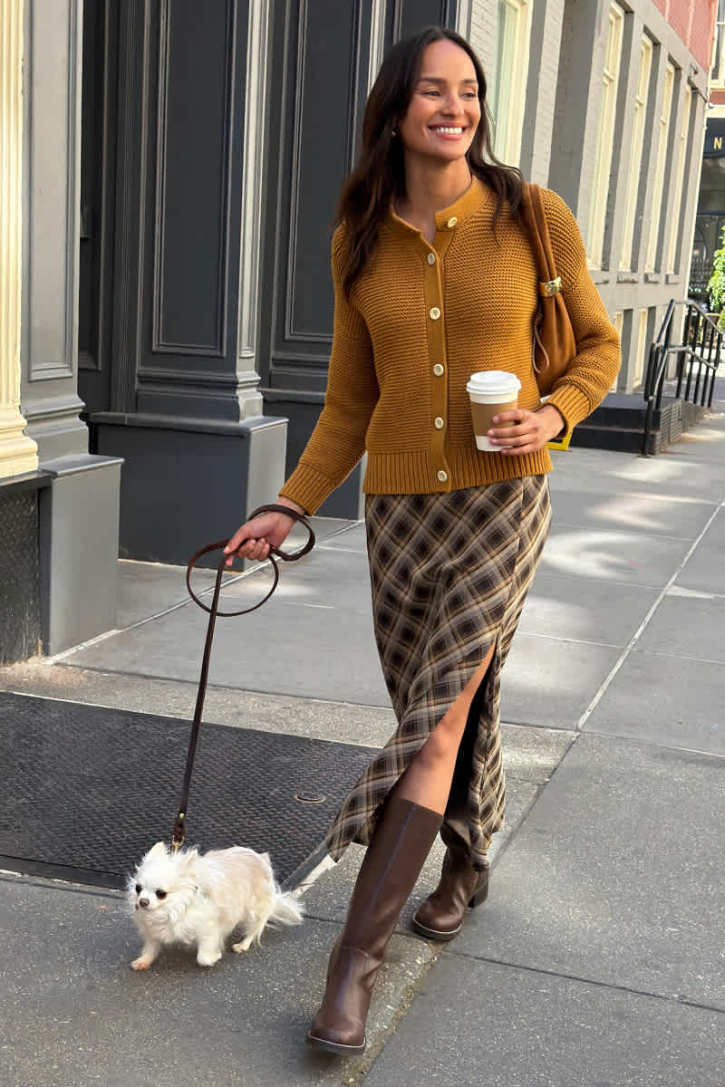 Model wearing Stitch Fix women's clothes including a mustard cardigan, a plaid midi skirt, dark brown knee-high boots, and a brown shoulder bag.