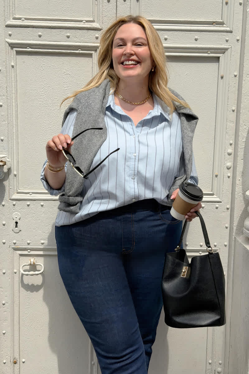 Model wearing Stitch Fix women's clothes including a light blue striped button-up shirt, dark denim jeans, a gray sweater, and a black handbag.