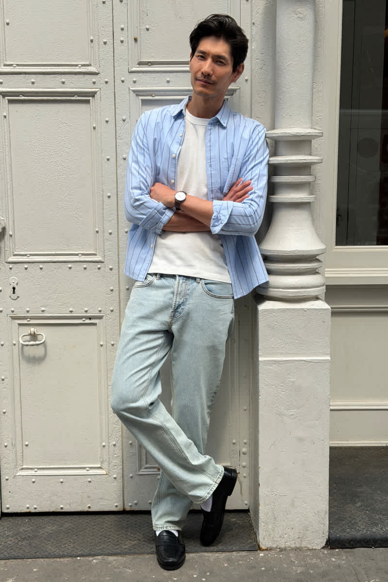 Model wearing Stitch Fix men's clothes including a light blue striped button-up shirt, a white T-shirt, light wash jeans, and black loafers.
